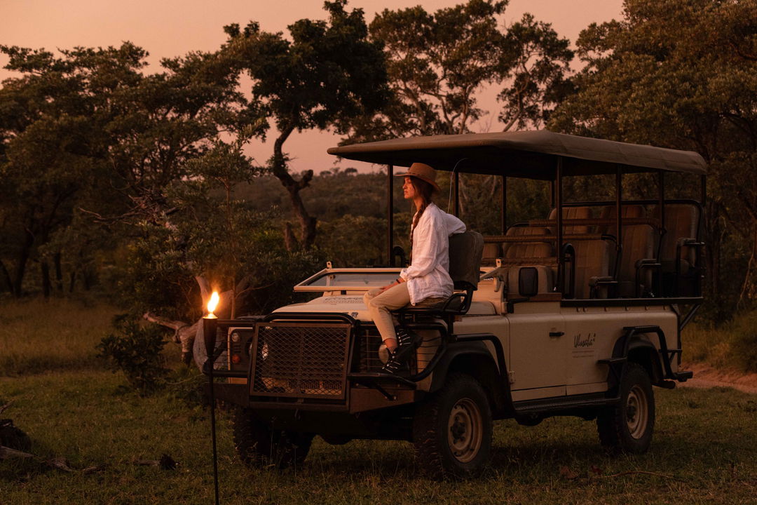 lady guest sitting on hood of safari vehicle in bush with fire in background at ulusaba safari lodge