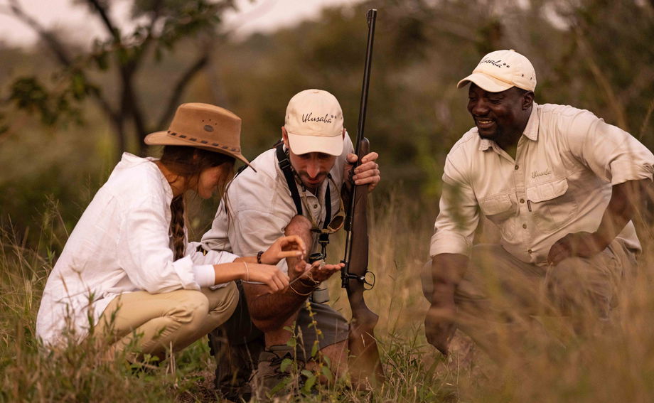 two safari bush guides kneeling in grass showing lady guests the tracks