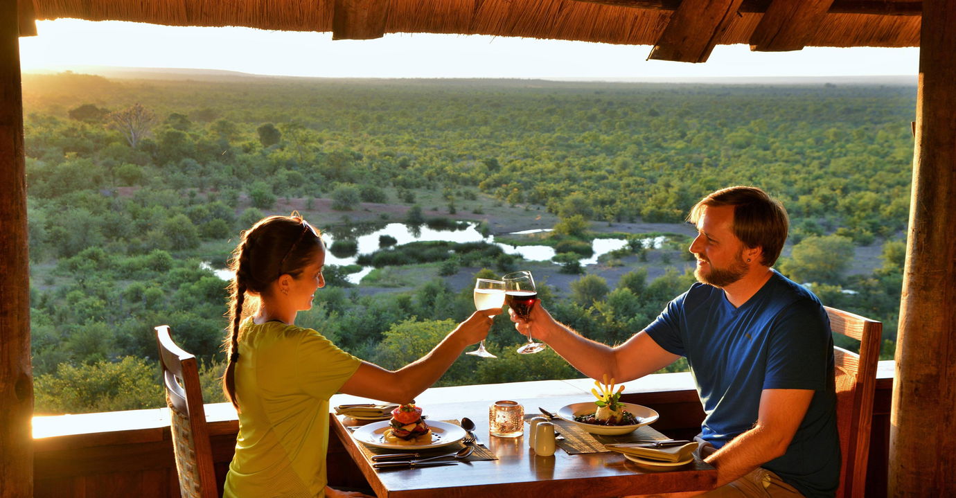 couple drinking wine at sunset dinner on deck of Victoria falls safari lodge with views of watering hole below 
