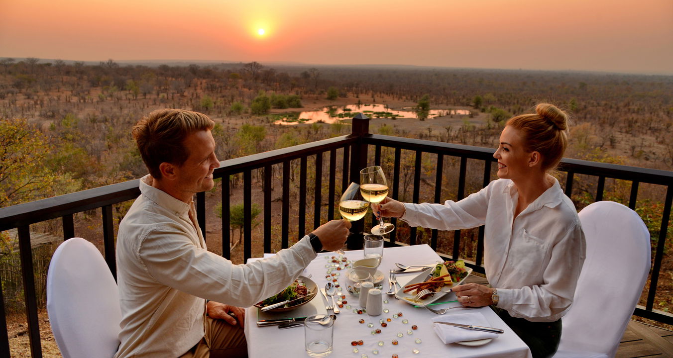 couple drinking wine at sunset dinner on deck of Victoria falls safari lodge with views of watering hole below 