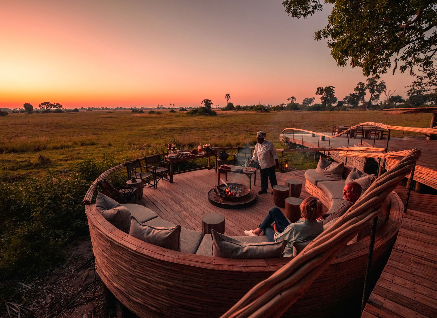 fire pit sunset view of deck and ring couches with staff preparing supper to two guests at Okavango Delta by Little Mombo Wilderness
