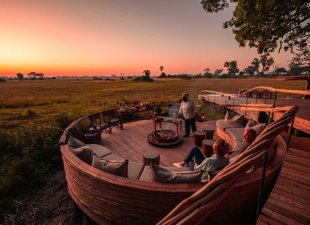 fire pit sunset view of deck and ring couches with staff preparing supper to two guests at Okavango Delta by Little Mombo Wilderness
