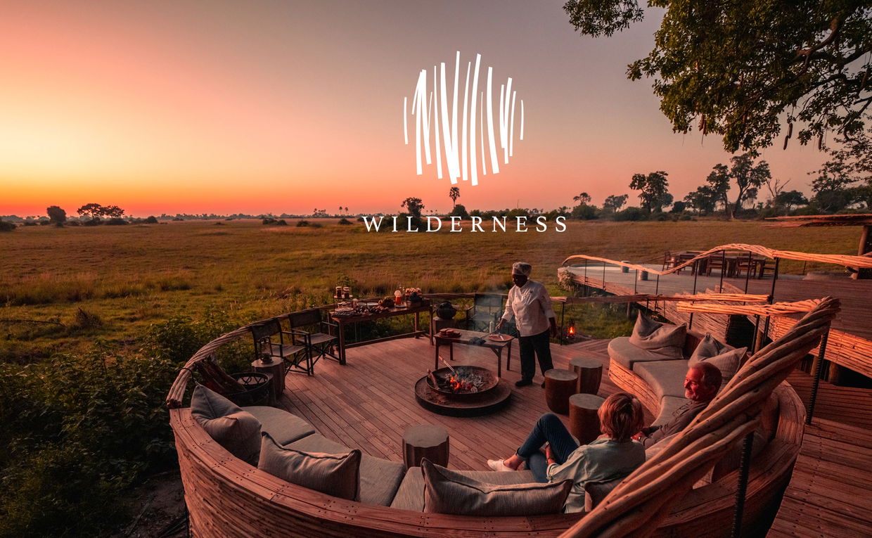 fire pit sunset view of deck and ring couches with staff preparing supper to two guests at Okavango Delta by Little Mombo Wilderness logo overlay fire pit sunset view of deck and ring couches with staff preparing supper to two guests at Okavango Delta by Little Mombo Wilderness logo overlay
