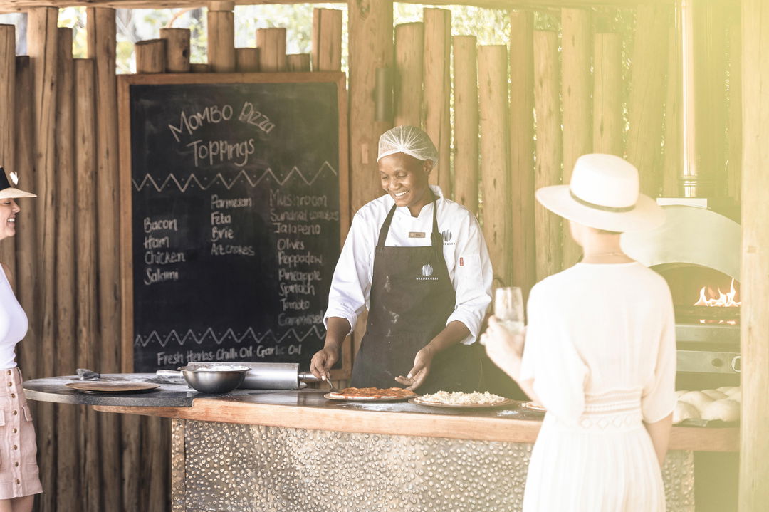 staff preparing pizza for guest in kitchen area of little mombo camp wilderness