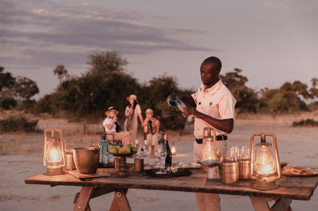 guide preparing sundowners on dressed table in bush for guests at Okavango Delta by Little Mombo Wilderness
