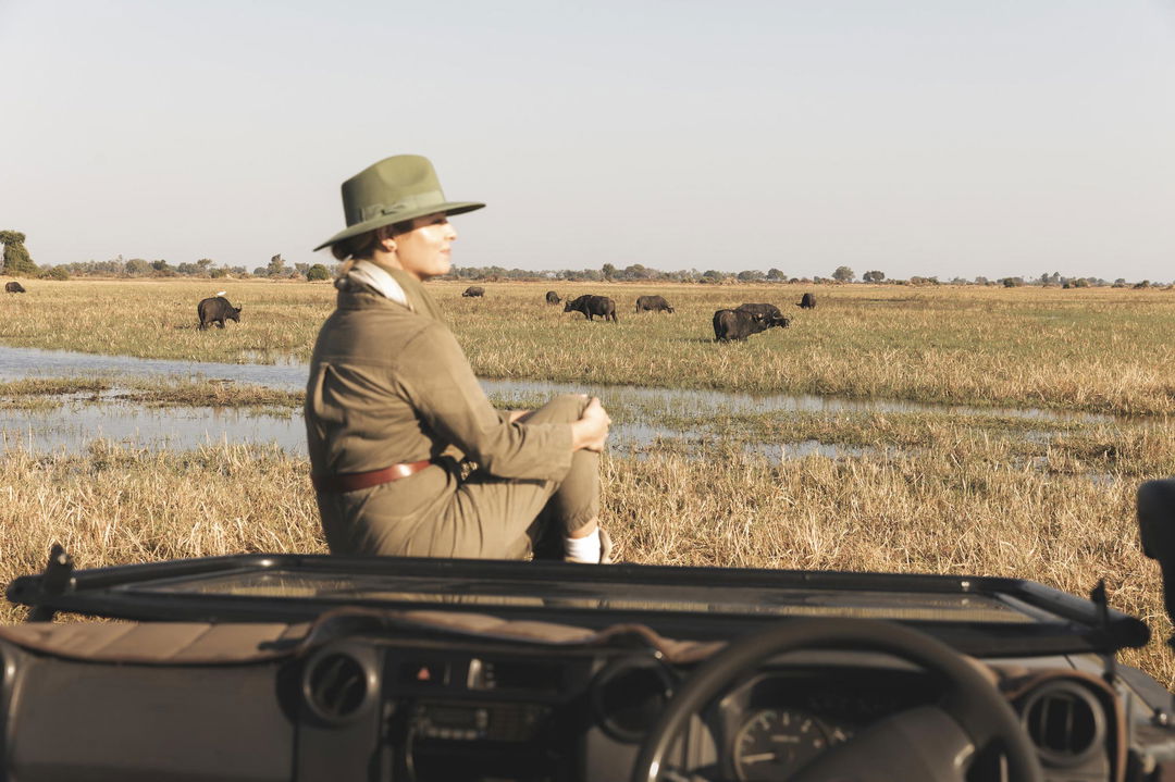 lady guest on hoof of safari vehicle looking at buffalo in the delta waters by Wilderness Mombo Camp Okavango Delta
