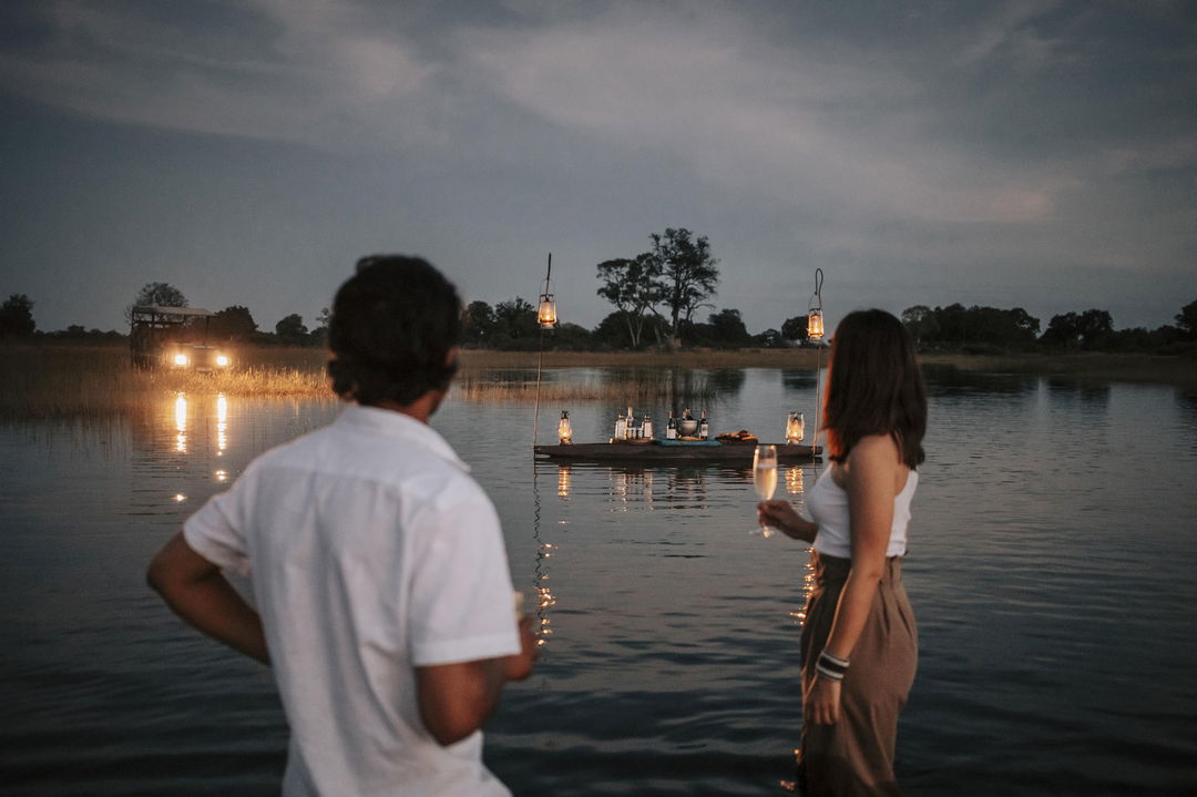 romantic dinner set up in water of delta with couple standing in water nearby at Vumbura Plains Camp Wilderness Okavango Delta